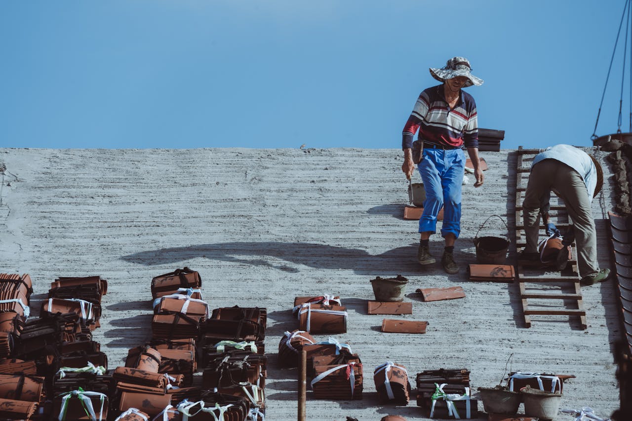 Two construction workers installing roof tiles on a building under a clear blue sky.